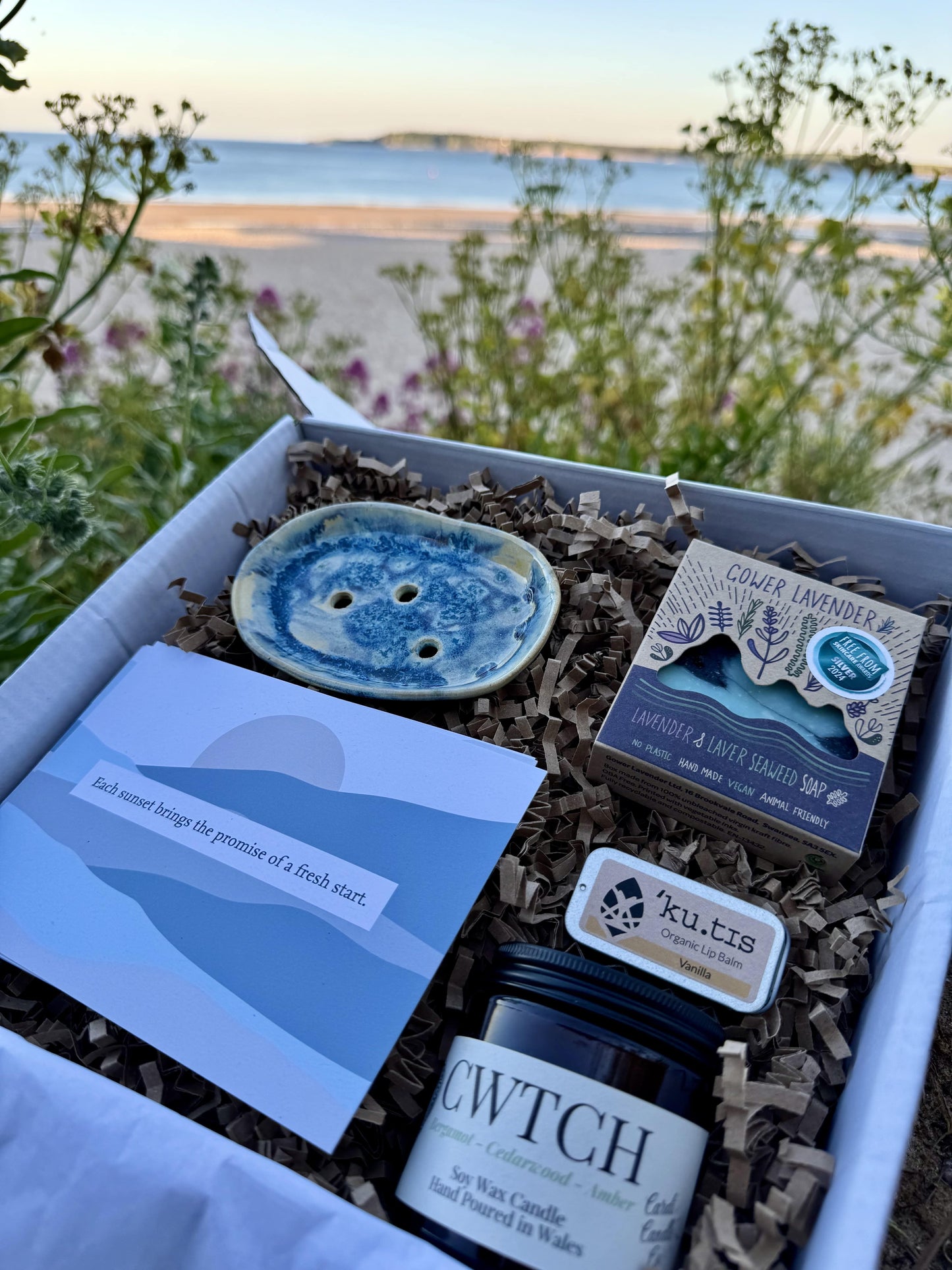 Wellness gift box by local Welsh makers, surrounded by sand and gentle waves in the background.