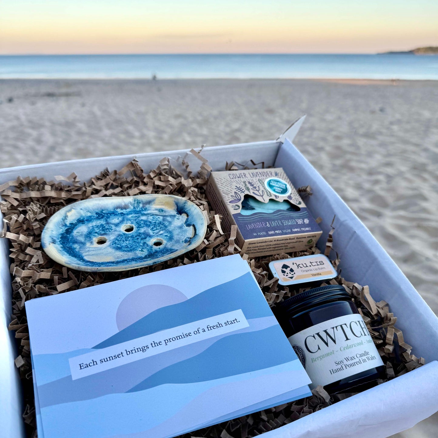 A wellness gift box with ceramic soap dish, soap, candle, lip balm and postcards, surrounded by sand and gentle waves in the background.