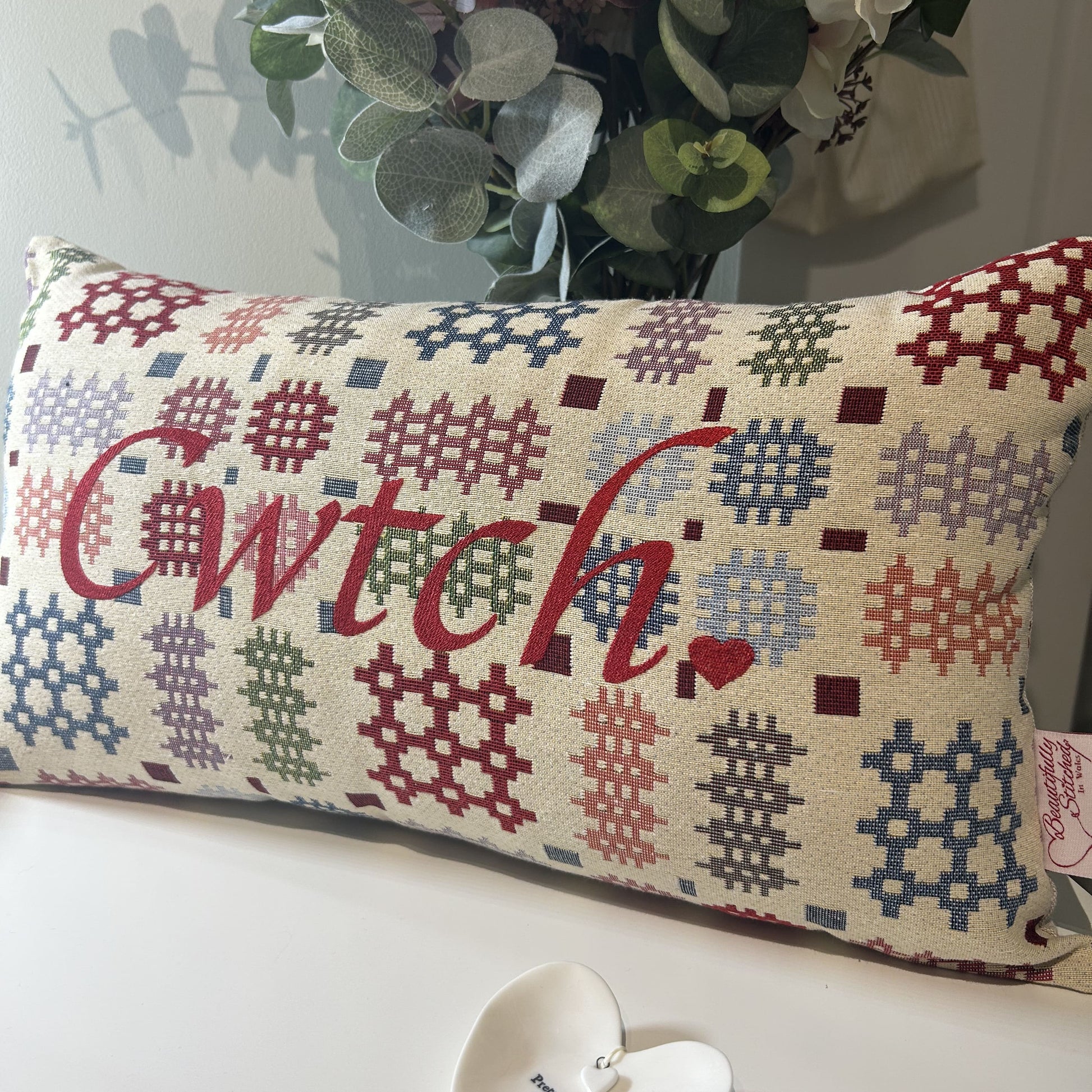 Decorative pillow with geometric pattern and 'Cwtch' text, placed on a surface with a heart-shaped object and plant in the background.