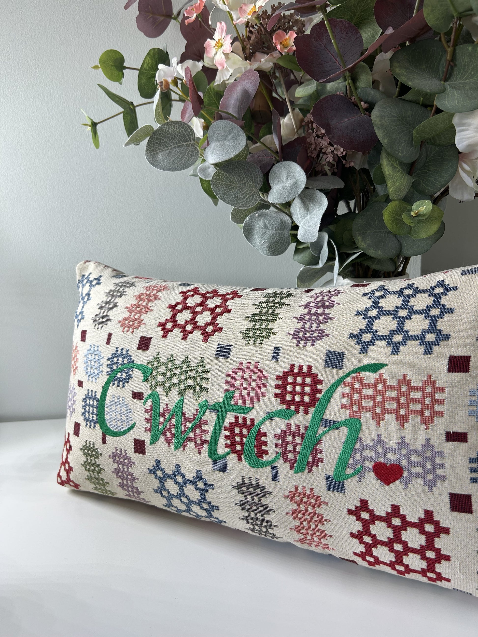 Decorative pillow with geometric pattern and 'Cwitch' text, placed on a surface with a plant in the background.