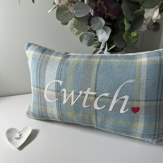 Plaid pillow with 'Cwtch' text and heart design on a surface with flowers in the background