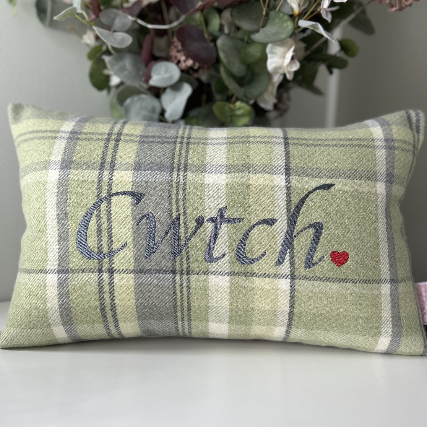 Plaid pillow with 'Cwtch' and a heart on a white surface with flowers in the background