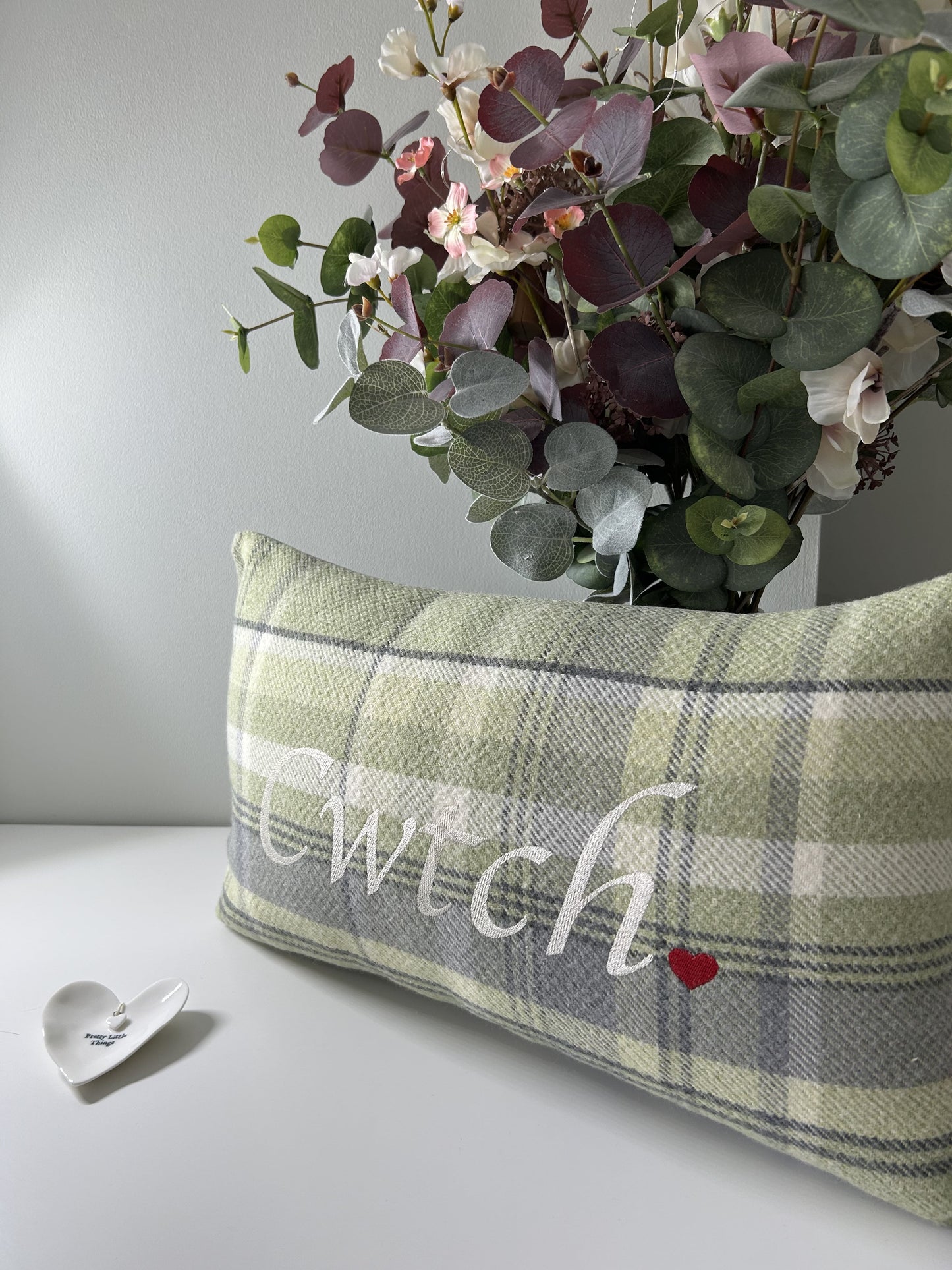 Plaid pillow with 'Wulch' embroidery on a white surface with flowers in the background