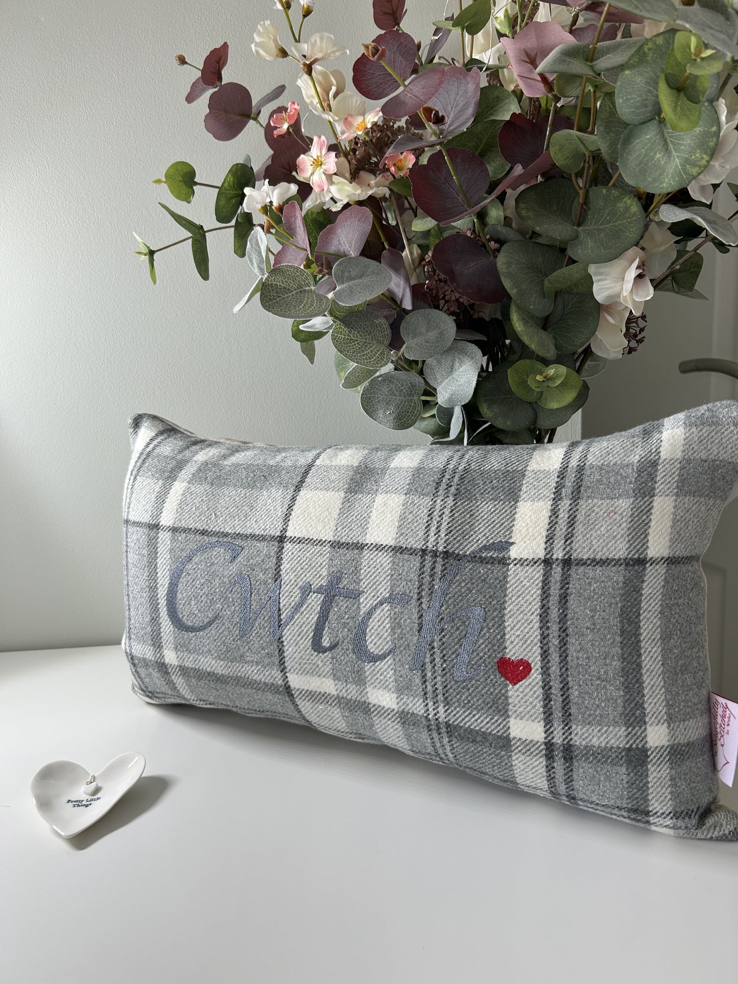 Plaid pillow with 'Cwtch' and a heart, accompanied by a bouquet of flowers on a light background.