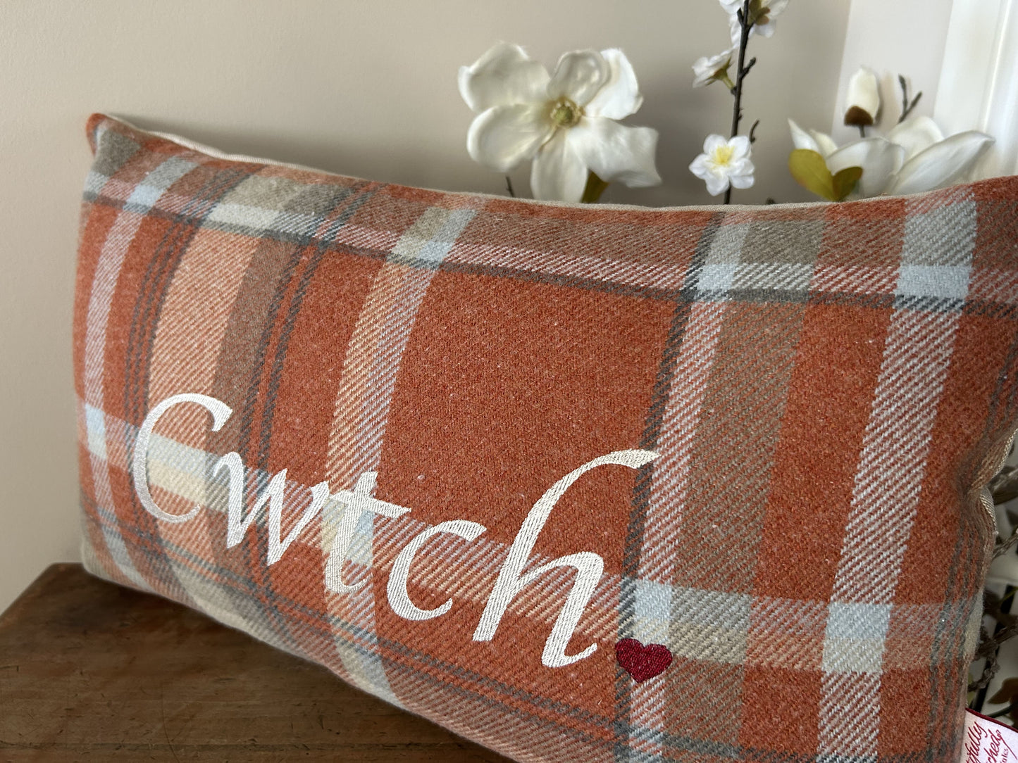 Plaid pillow with 'Cwtch' and a heart on a wooden surface with flowers in the background