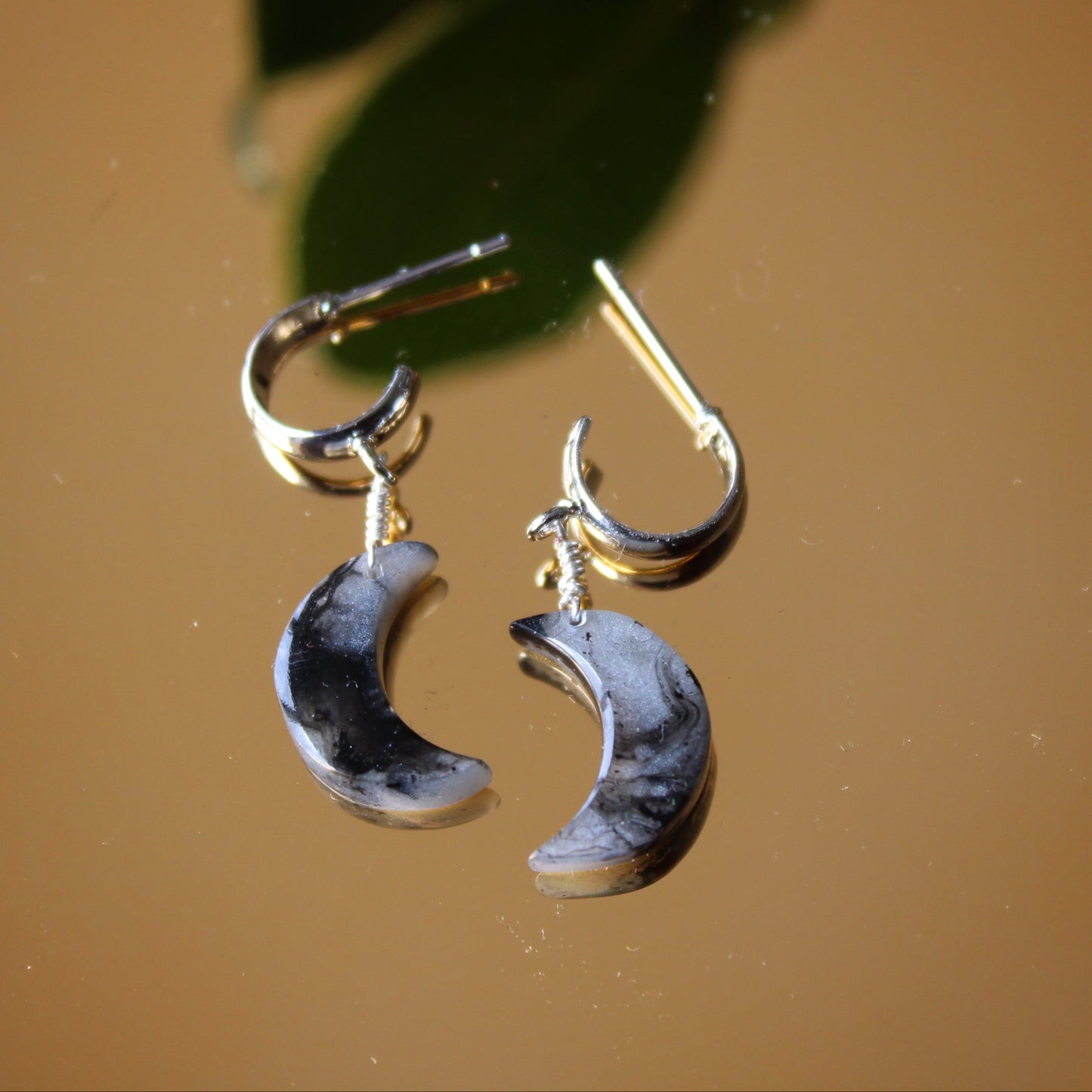 Crescent moon-shaped earrings on a reflective surface with green leaves in the background