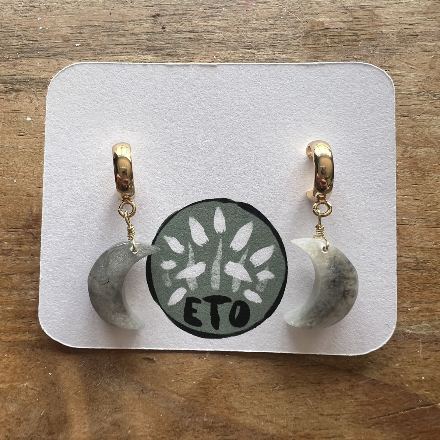 Pair of earrings on a display card with 'ETO' branding on a wooden surface, elegantly crafted for a unique accessory choice.