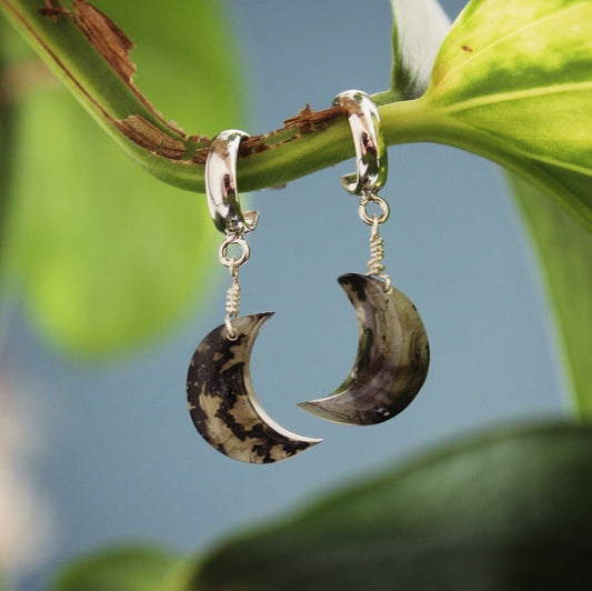Moon-shaped earrings hanging from a leaf with a blurred green background, elegantly crafted for a unique accessory choice.