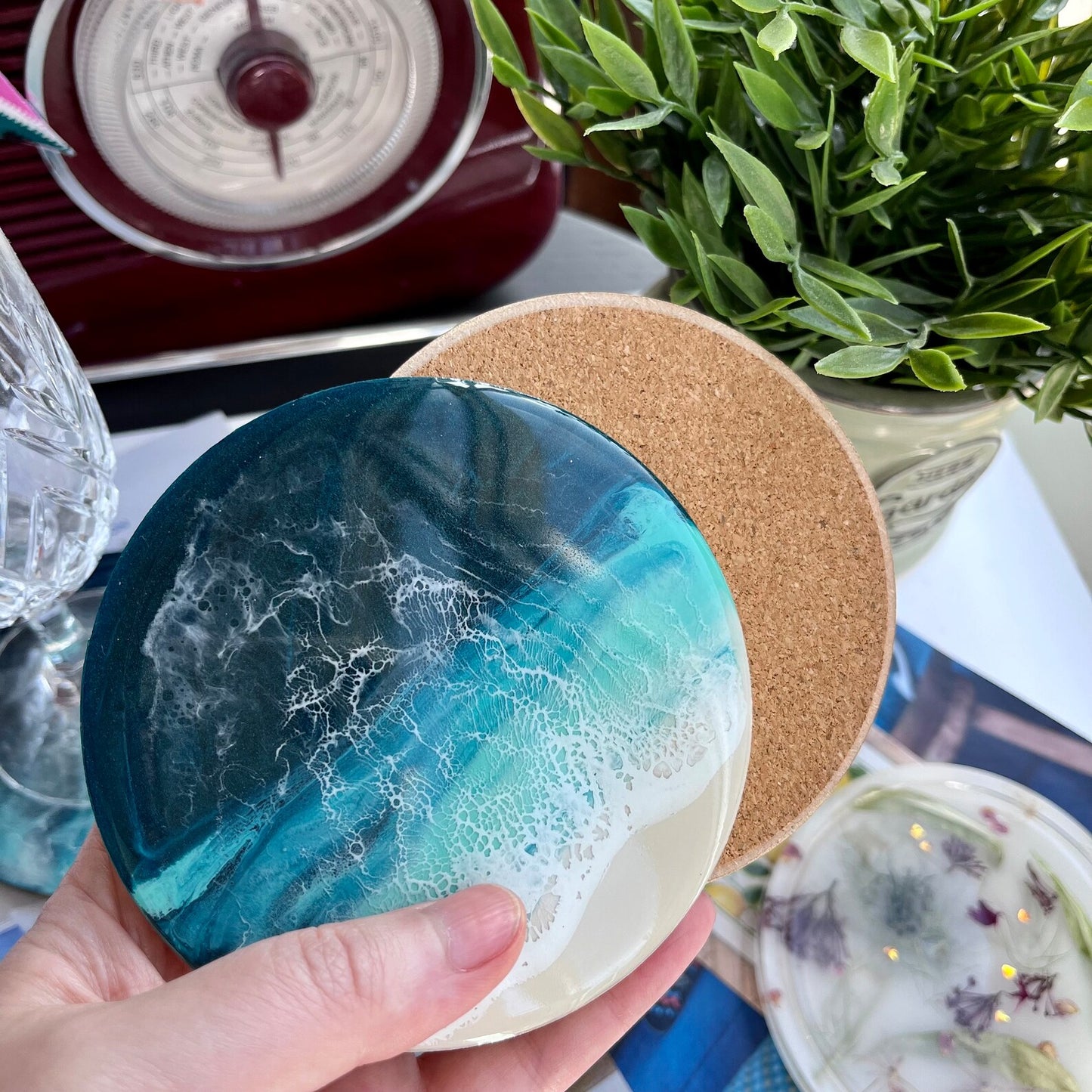 Coaster with blue and white design held by a hand, with a plant and decorative items in the background.