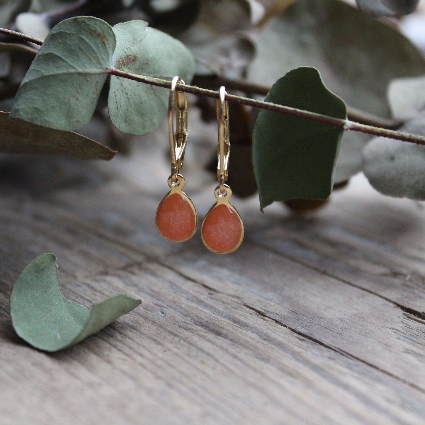 Oval drop shaped earrings with gold hooks on a wooden surface with eucalyptus leaves.