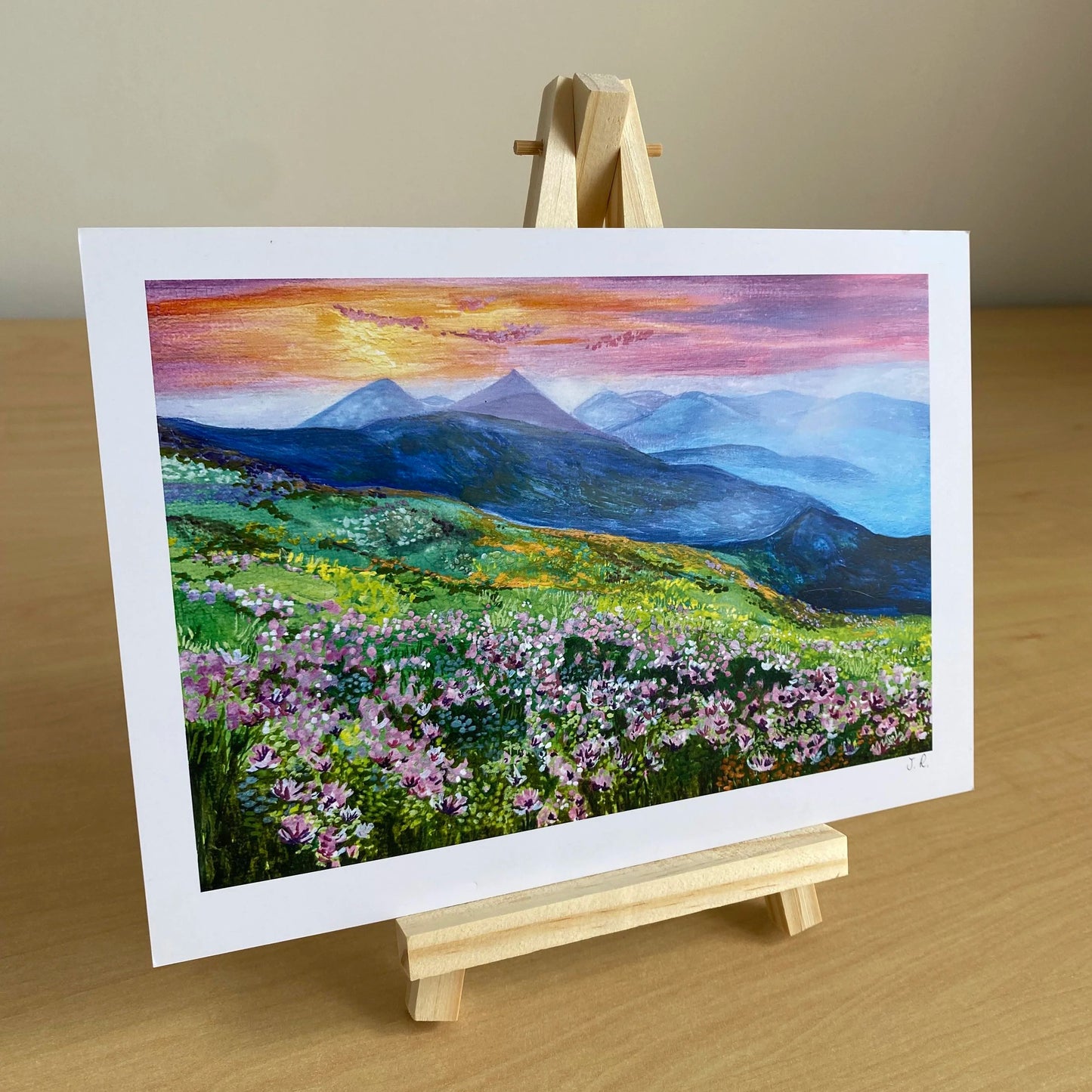 Painting of a mountain landscape with flowers on an easel against a beige wall. Sunset landscape print with floral foreground for cosy interiors.