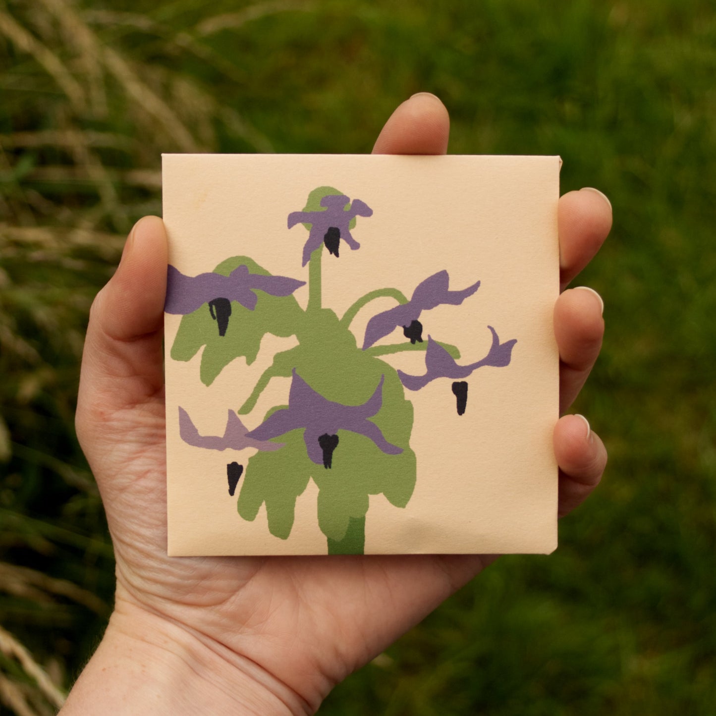 Hand holding a square beige card with green and purple floral design against a blurred natural background