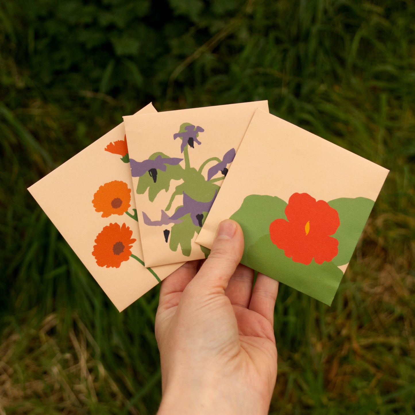 Hand holding three small envelopes with floral designs against a grassy background