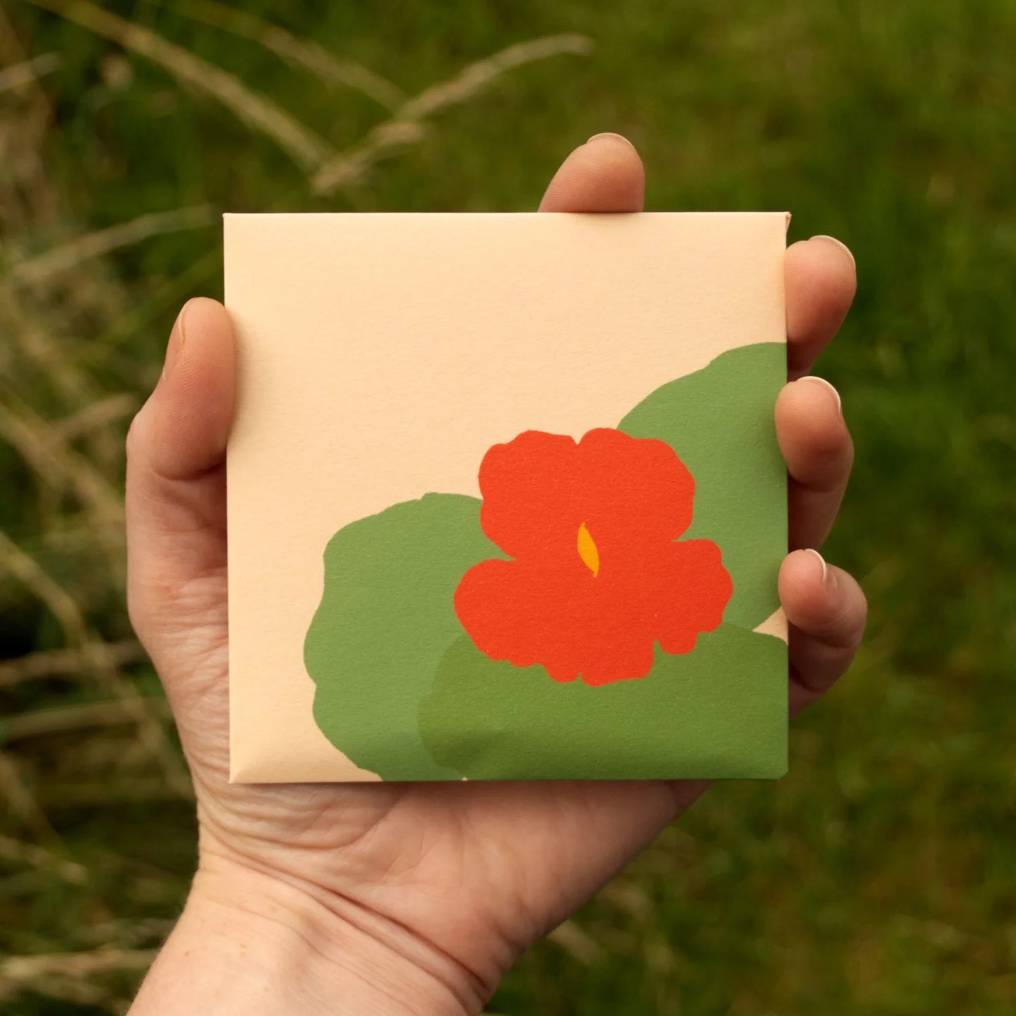 Hand holding a small square box with a red flower design against a natural background