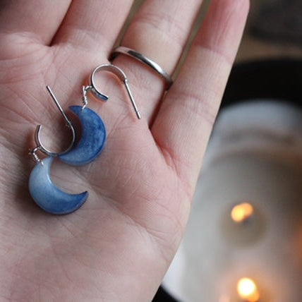 Blue handmade resin moon-shaped earrings held in a hand with a blurred candlelight background. Made in Wales. Perfect christmas gift for her.