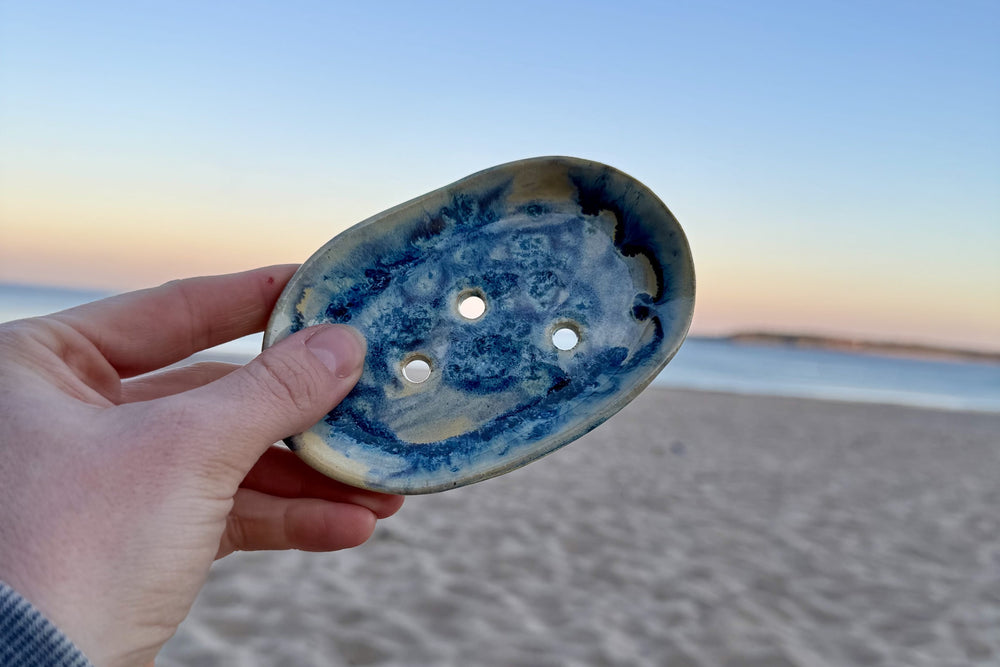 Ceramic soap dish by local Welsh maker, surrounded by sand and gentle waves in the background.