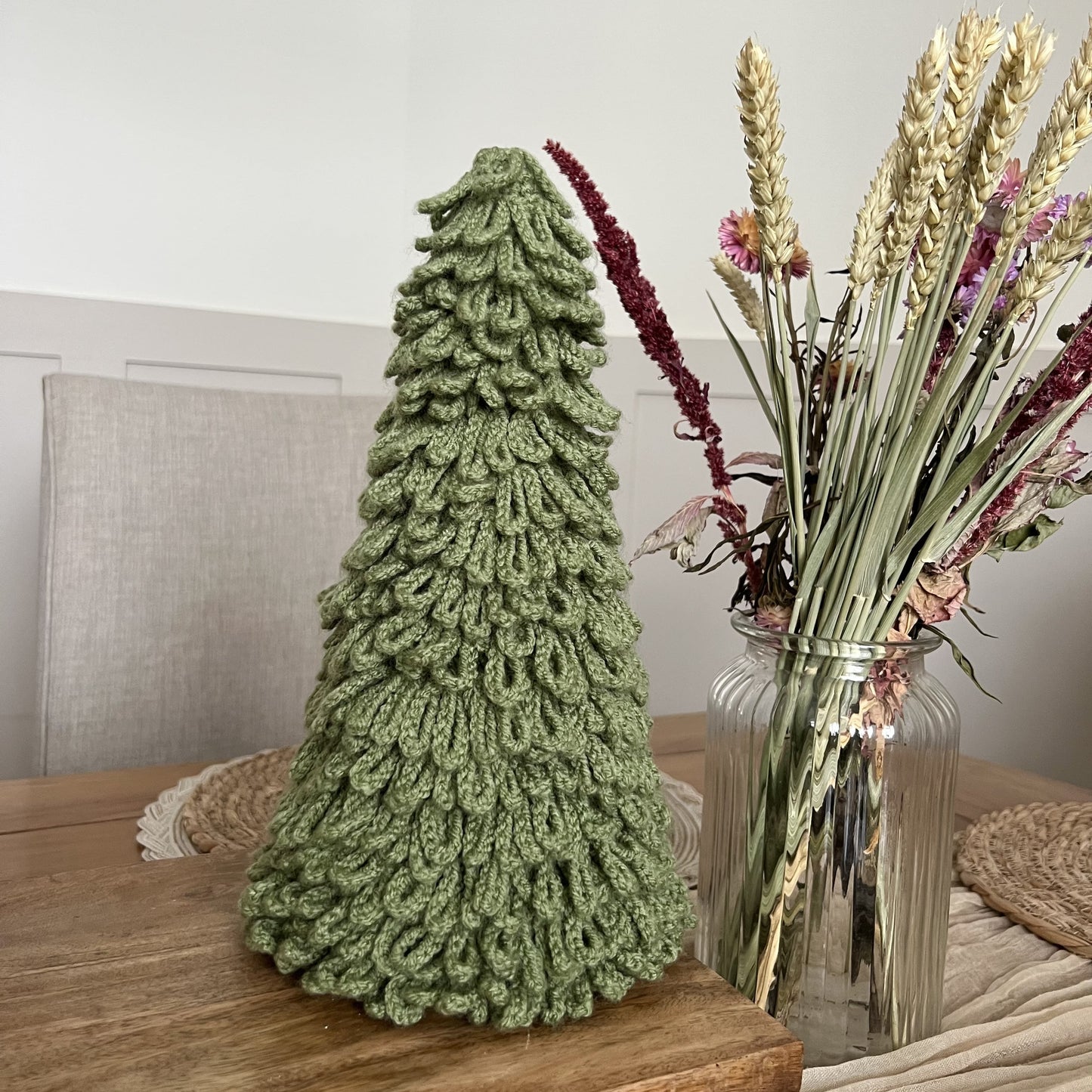 Crocheted loopy Christmas tree decor in soft sage yarn, handmade in Wales, on a wooden surface with a vase of flowers in the background.