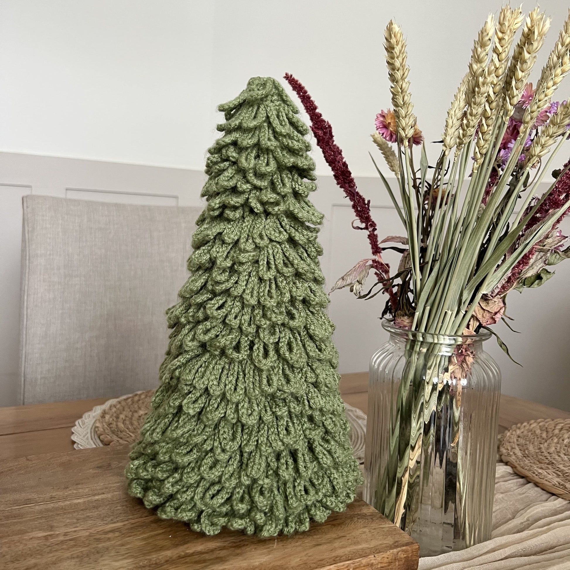 Crocheted loopy Christmas tree decor in soft sage yarn, handmade in Wales, on a wooden surface with a vase of flowers in the background.