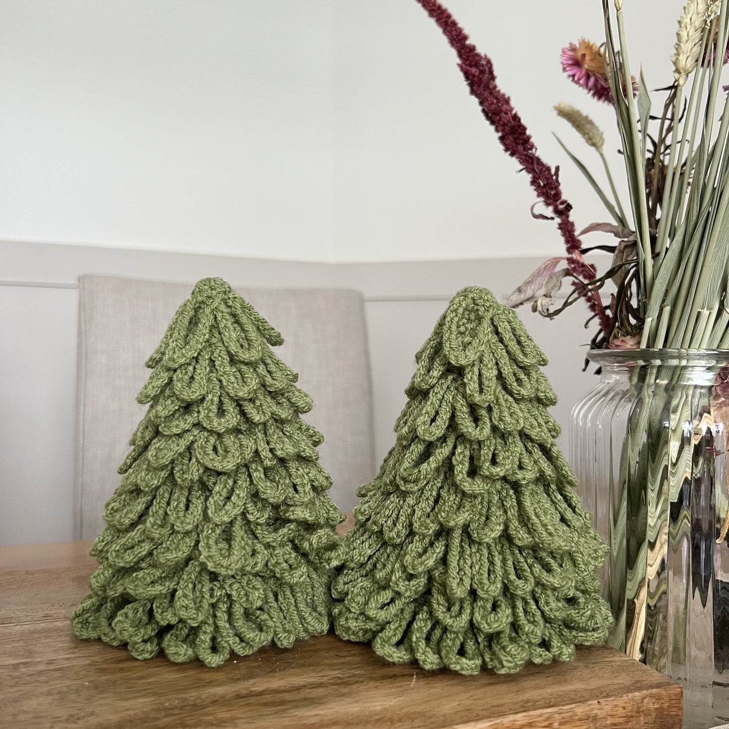 Two sage green crochet Christmas tree  decorations for home decor, displayed on a wooden table with a vase of flowers in the background.