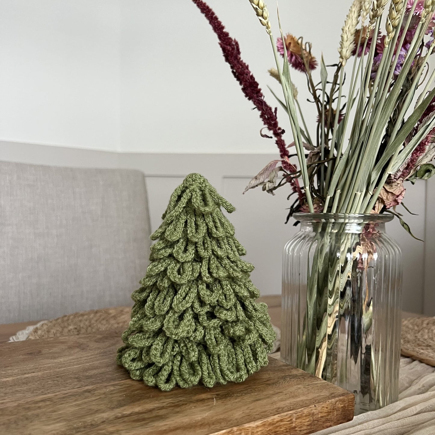 Decorative handmade crochete sage green Christmas tree and vase with flowers on a wooden surface. 