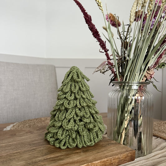 Decorative handmade crochete sage green Christmas tree and vase with flowers on a wooden surface. 