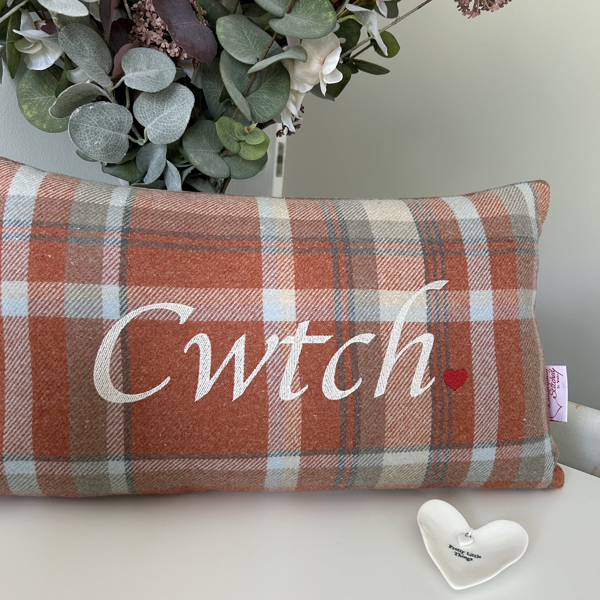 Plaid pillow with 'Cwtch' text and heart symbol on a surface with eucalyptus leaves in the background.
