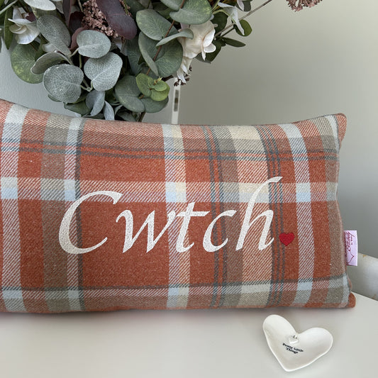 Plaid pillow with 'Cwtch' text and heart symbol on a surface with eucalyptus leaves in the background.
