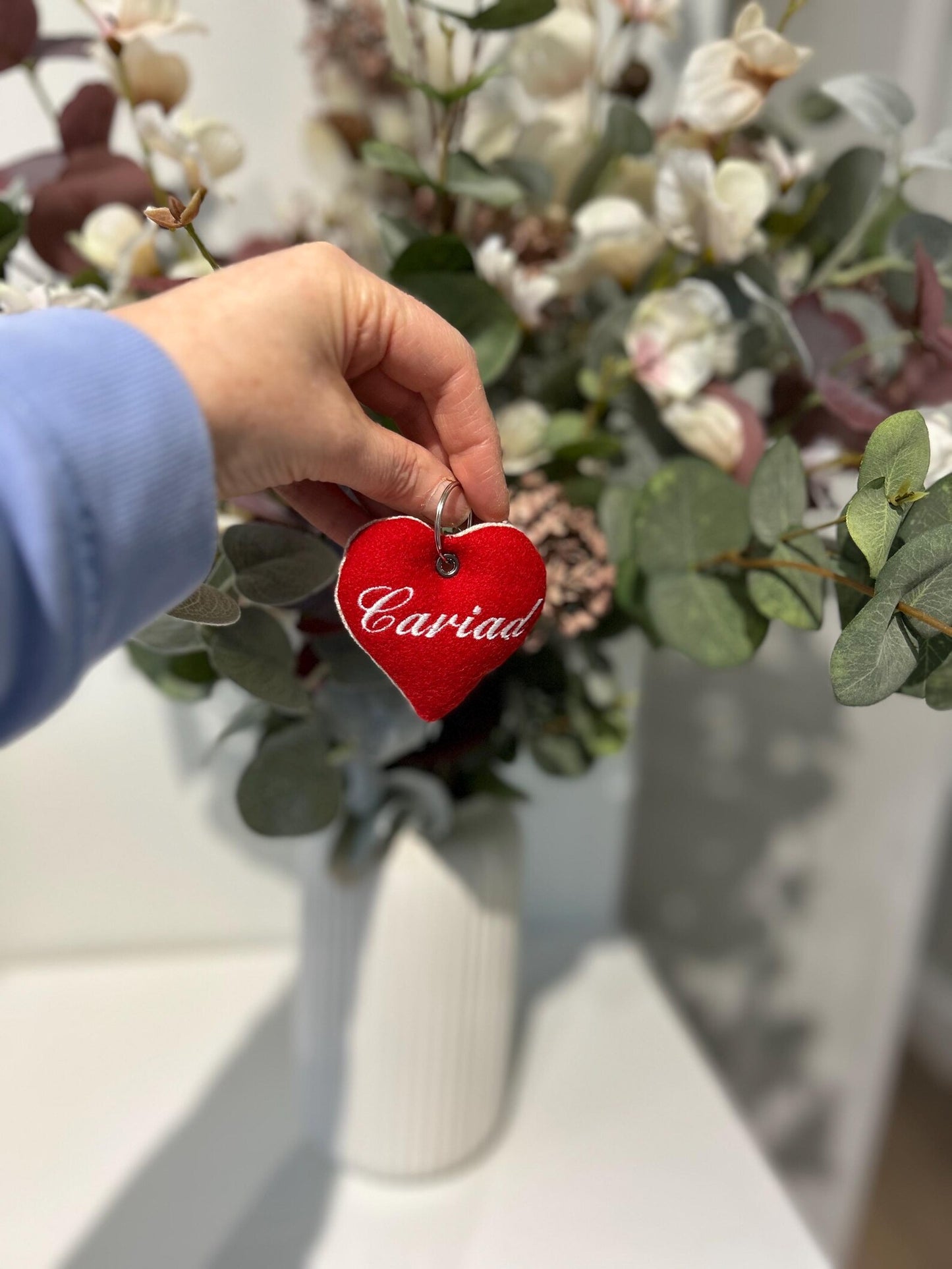 Hand holding a red handmade heart-shaped 'Cariad' keyring in front of a floral arrangement. Embroidered with “Cwtch” in red thread, unique Welsh textile gift accessory for birthdays, Christmas, weddings