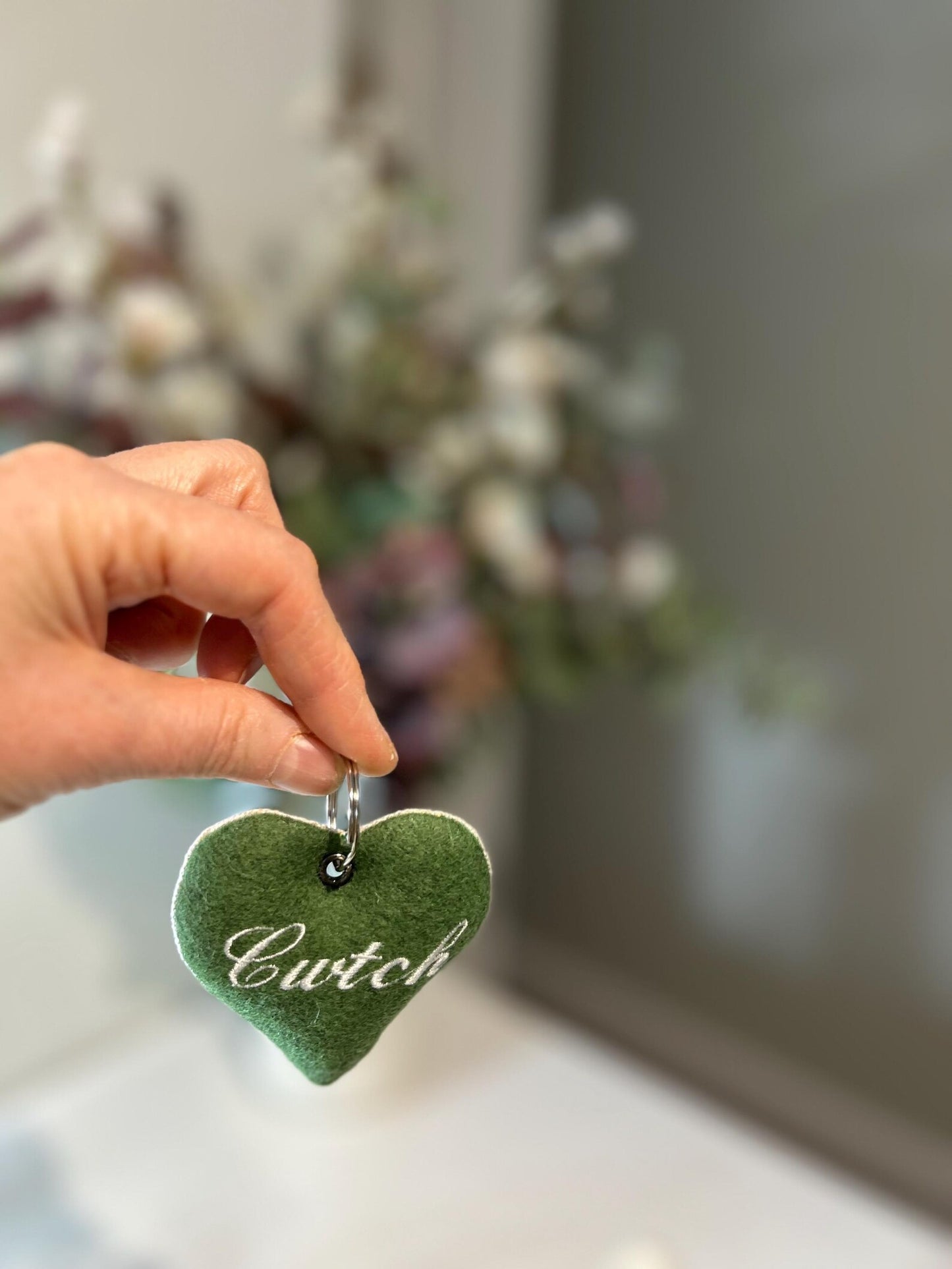 Green heart-shaped handmade keyring with 'Cwtch' text held by a hand against a blurred indoor background. unique Welsh textile gift accessory for birthdays, Christmas, weddings