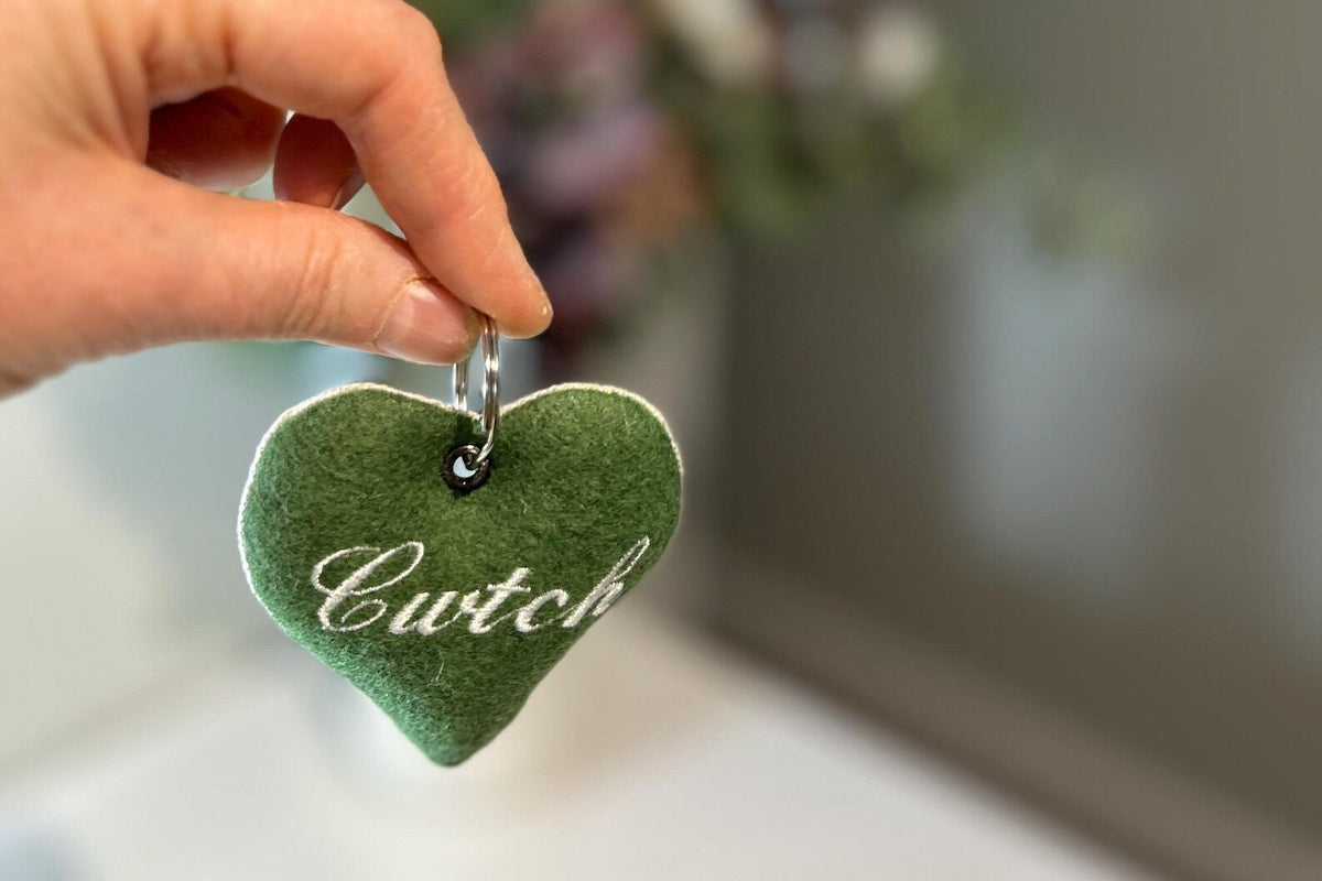 Green heart-shaped handmade keyring with 'Cwtch' text held by a hand against a blurred indoor background. unique Welsh textile gift accessory for birthdays, Christmas, weddings