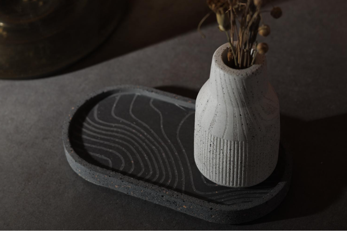 A small ribbed ceramic vase in off-white sits on a dark oval tray, holding dried botanical stems. The scene is styled with warm ambient lighting and blurred dried flowers in the background.