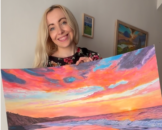 A smiling blonde woman holds up a large canvas painting of a colourful sunset beach scene with pink, orange, purple, and blue tones, photographed indoors in a home setting.