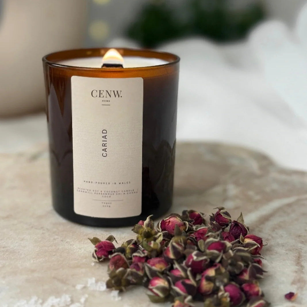 Cariad peony and suede soy candle hand-poured in Wales, with a label on a surface with dried flowers. 