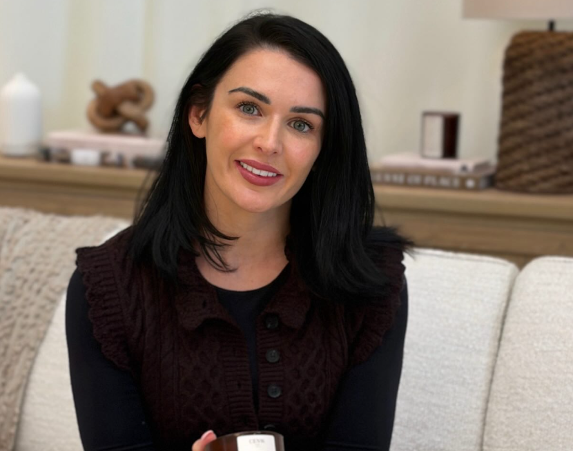 A woman sitting on a light beige couch holds a dark amber glass jar candle with a white label. She is wearing a black long-sleeve top and a dark brown cable-knit vest.