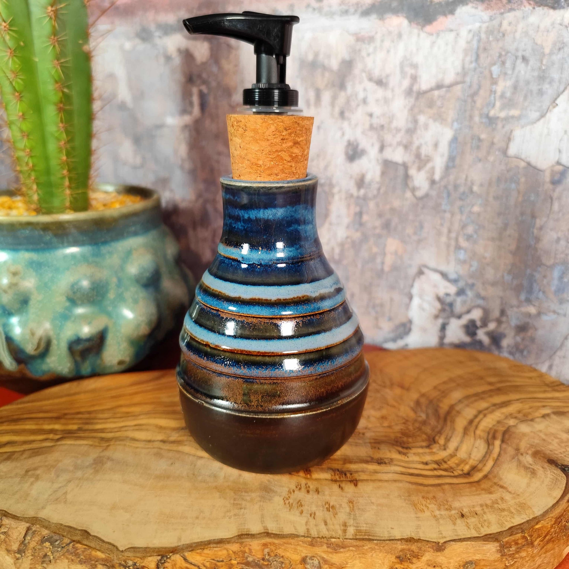 Ceramic soap dispenser with pump on a wooden surface next to a cactus plant. Handmade ceramic soap dispenser for bathroom or kitchen