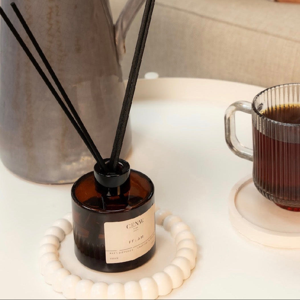 A diffuser with reeds on a table next to a mug of coffee and a vase. 100ml made in Wales with natural fragrance oils.