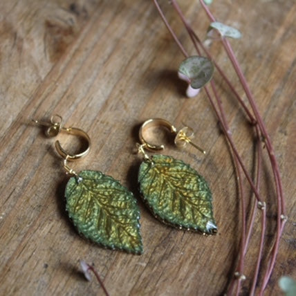 Green and gold leaf huggie earrings handmade resin jewellery on wooden surface with dried flowers.