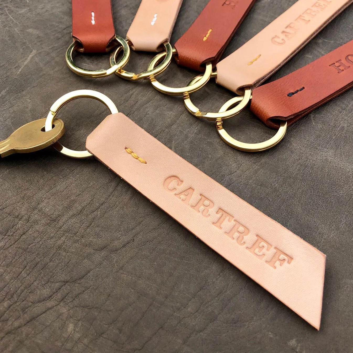 Leather keychains with gold rings on a dark surface, featuring the brand 'Cartref'. Handmade leather keyring with Cartref Welsh wording,