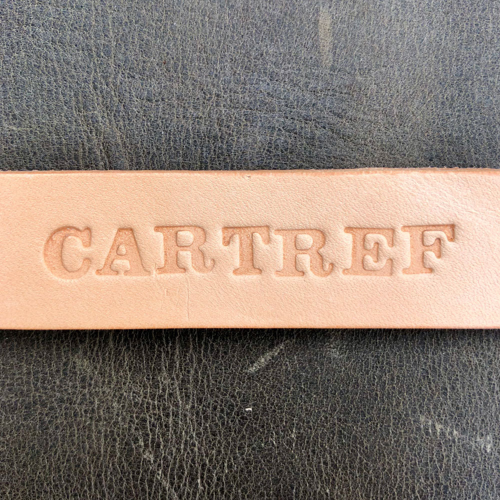 Close-up of a leather strap with 'CARTREF' embossed on it, against a textured gray background. Handmade leather keyring with Cartref Welsh wording