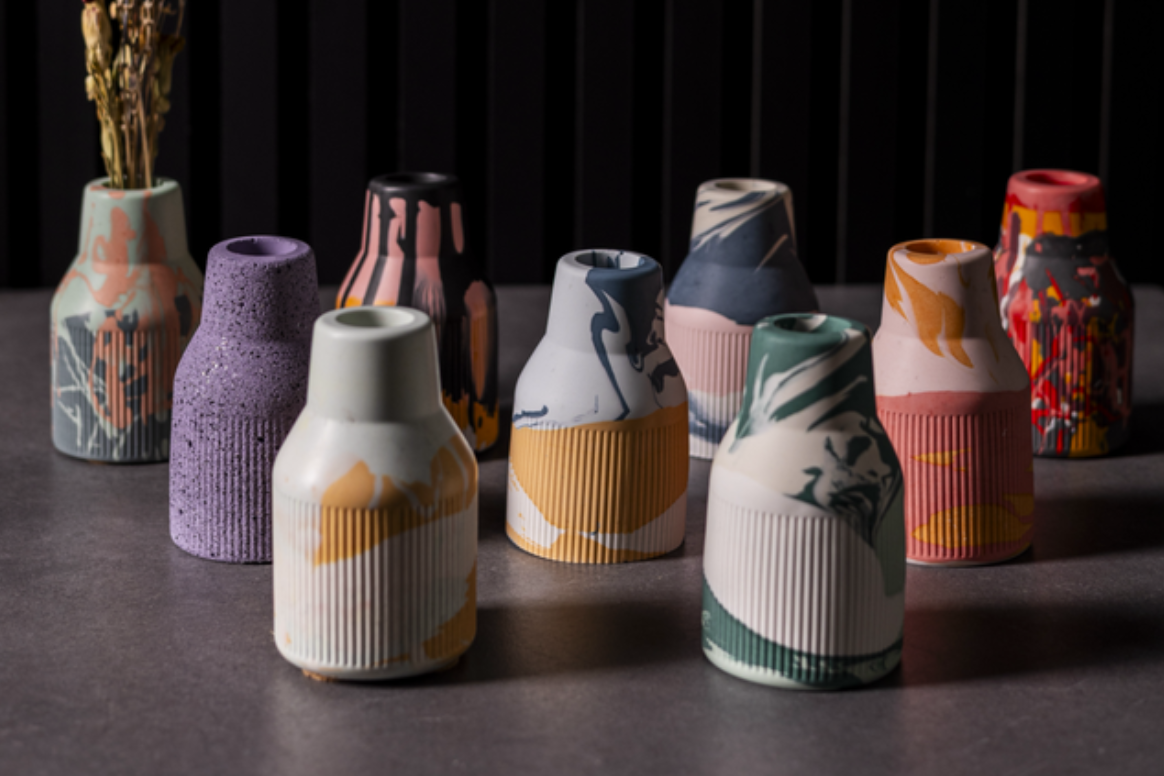 A group of decorative ceramic vases in assorted colors and patterns, including marble, speckled, and abstract designs, arranged on a dark tabletop with a black slatted background.