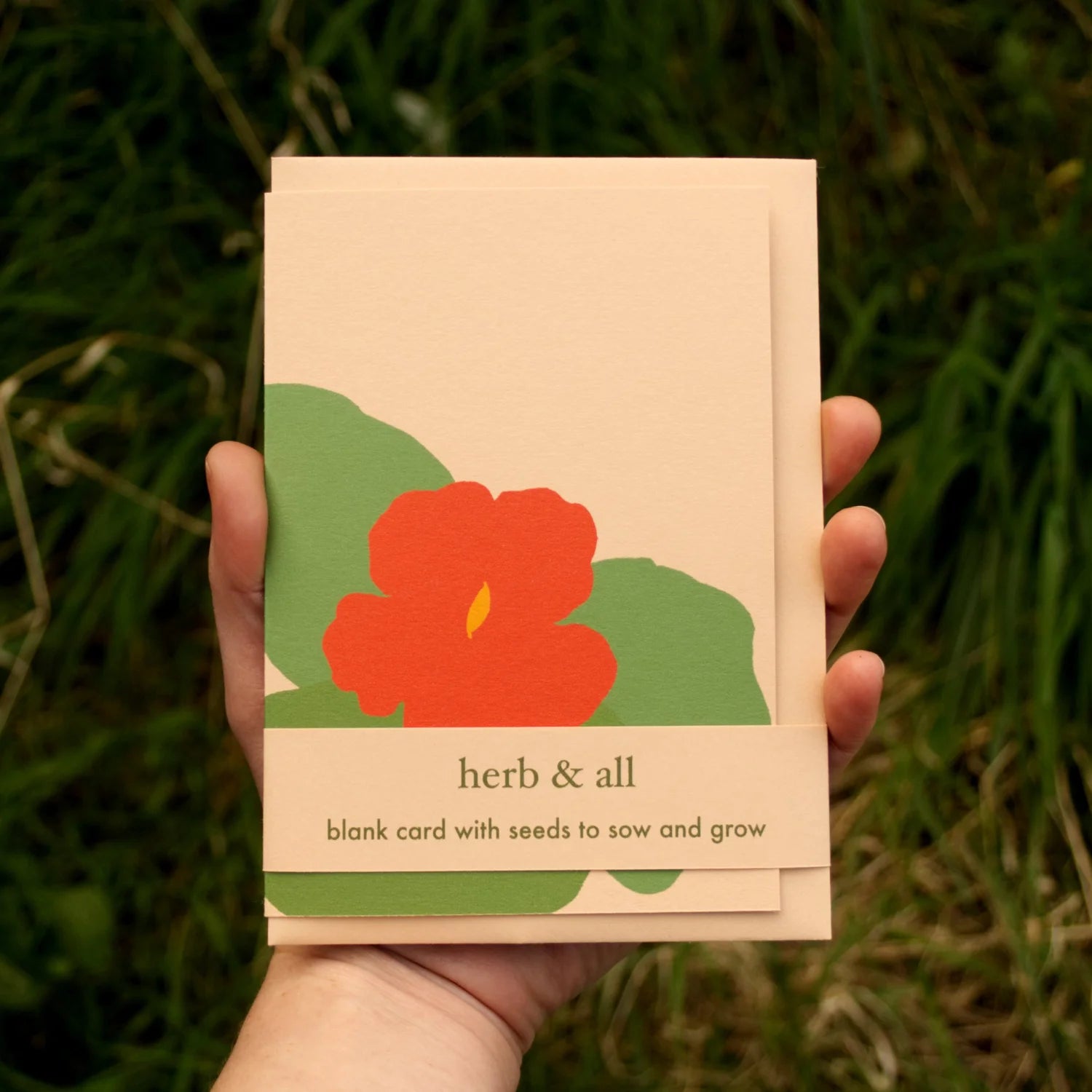Hand holding a card with a red flower design and text 'herb & all' against a green grass background.