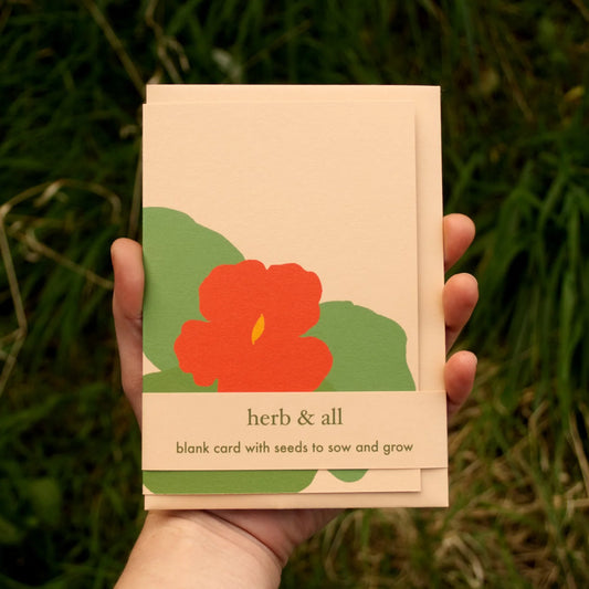 Hand holding a card with a red flower design and text 'herb & all' against a green grass background.