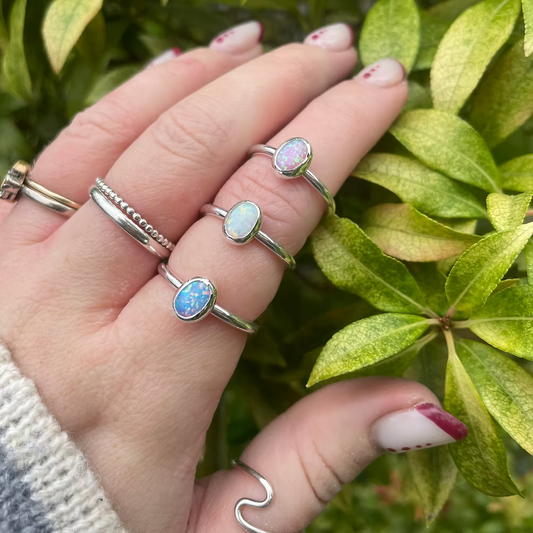 Hand wearing multiple silver rings with opal stones against a green leafy background. lab-grown opal ring, 8x6mm stone set in sterling silver,  made in Wales.