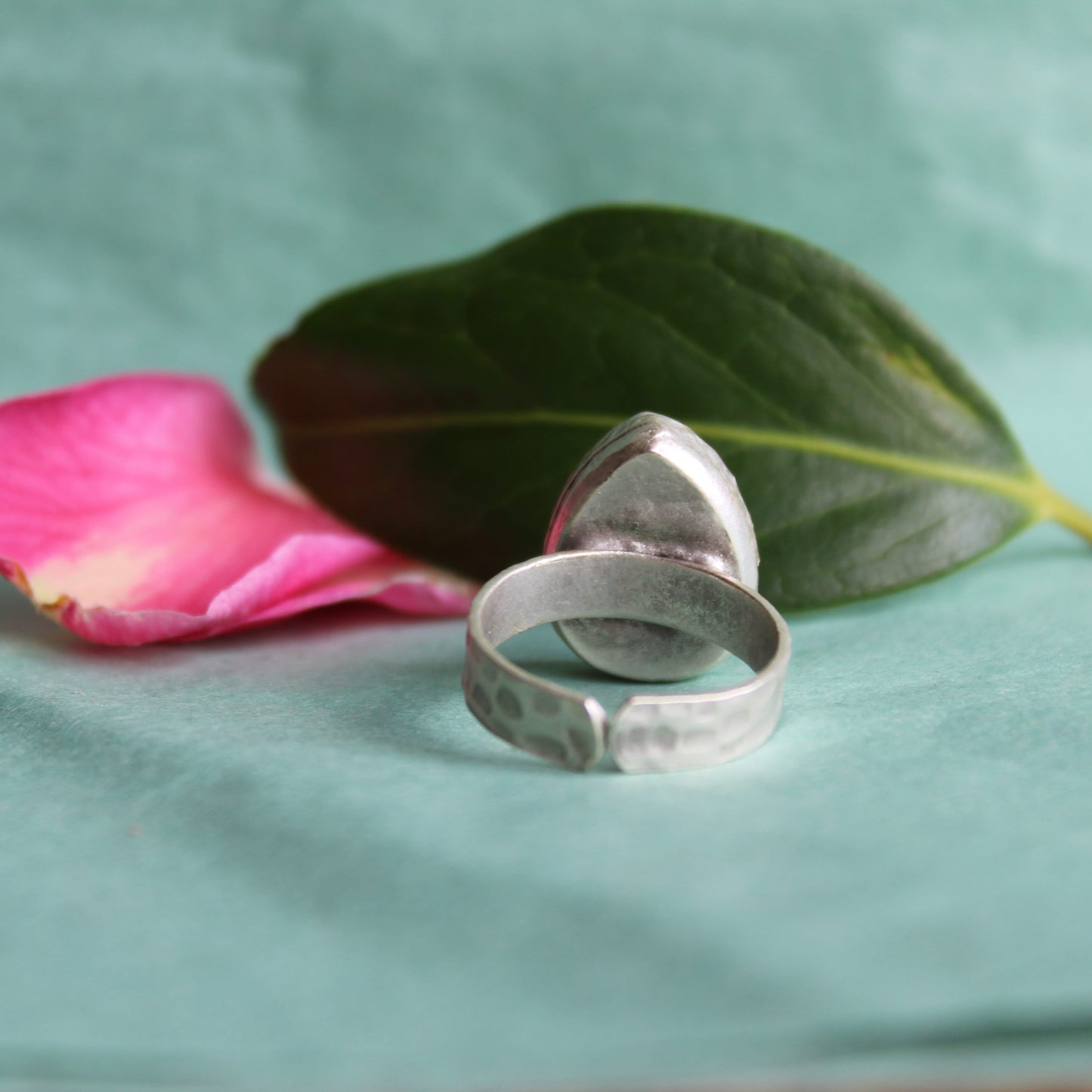 Silver ring on a teal surface with a pink rose petal and green leaf. Small business in Wales.