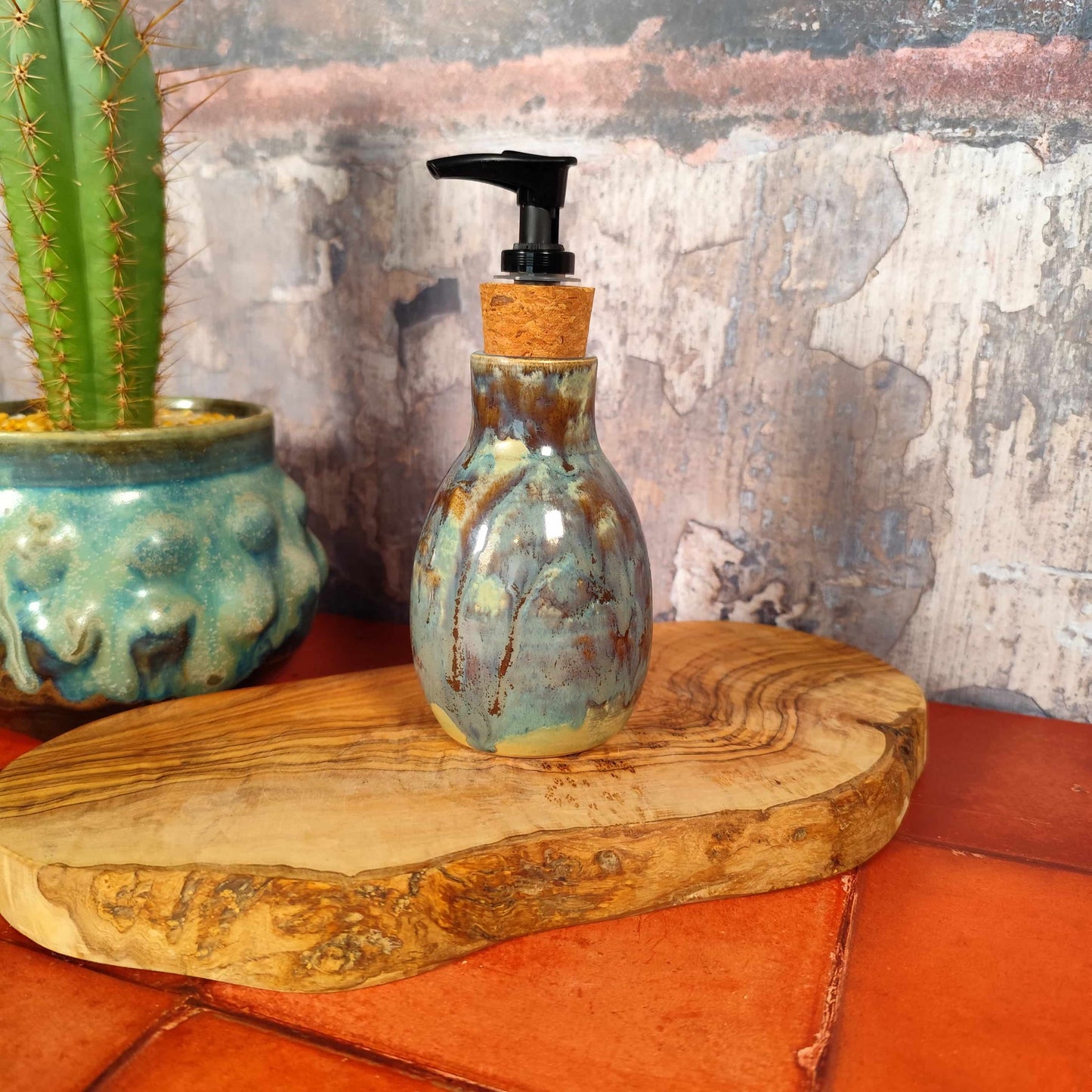 Handmade ceramic bottle with pump in cerulean and olive glaze, made in Wales. On a wooden surface next to a cactus plant. 