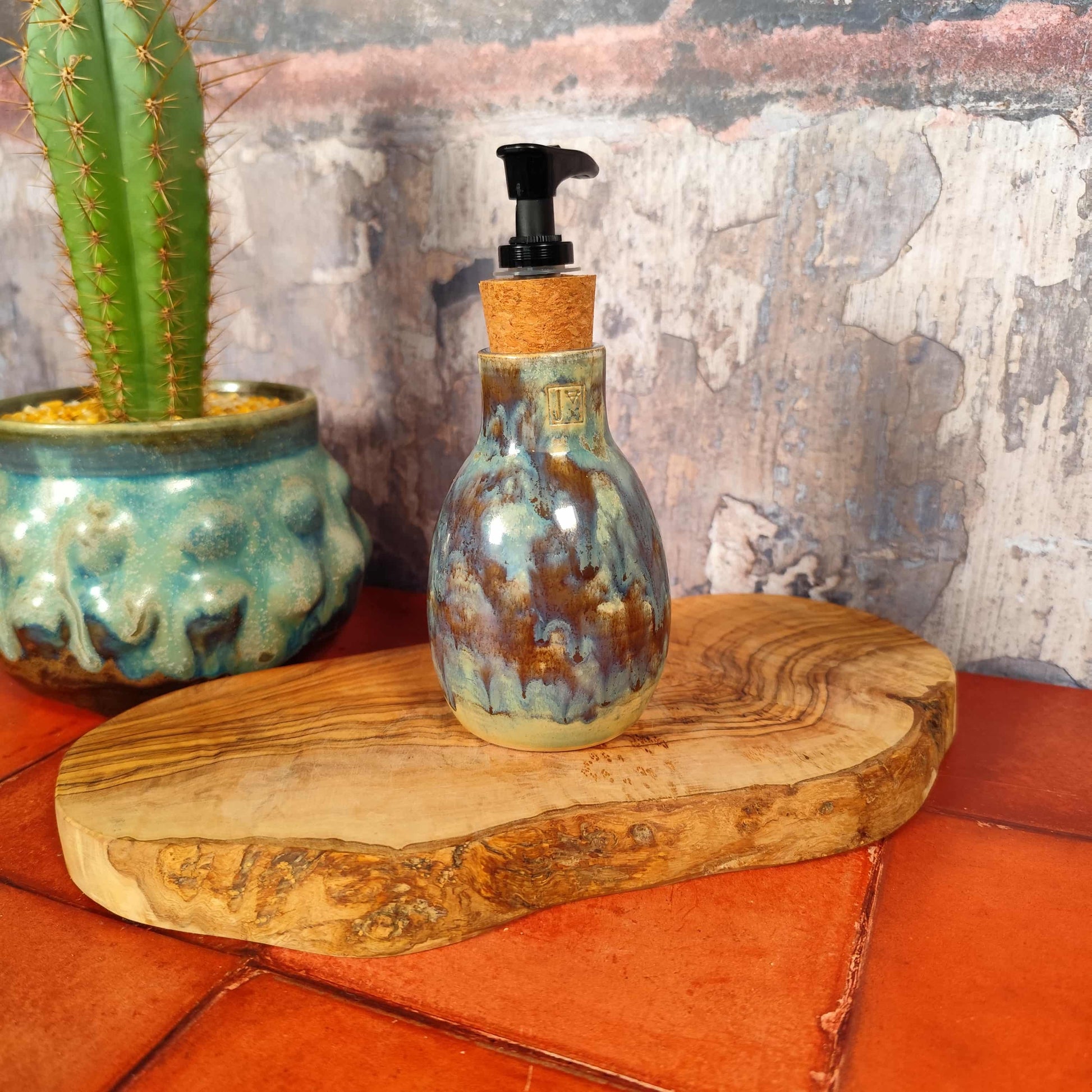 Handmade ceramic bathroom soap dispenser in cerulean and olive glaze, made in Wales. On a wooden surface next to a cactus plant. 