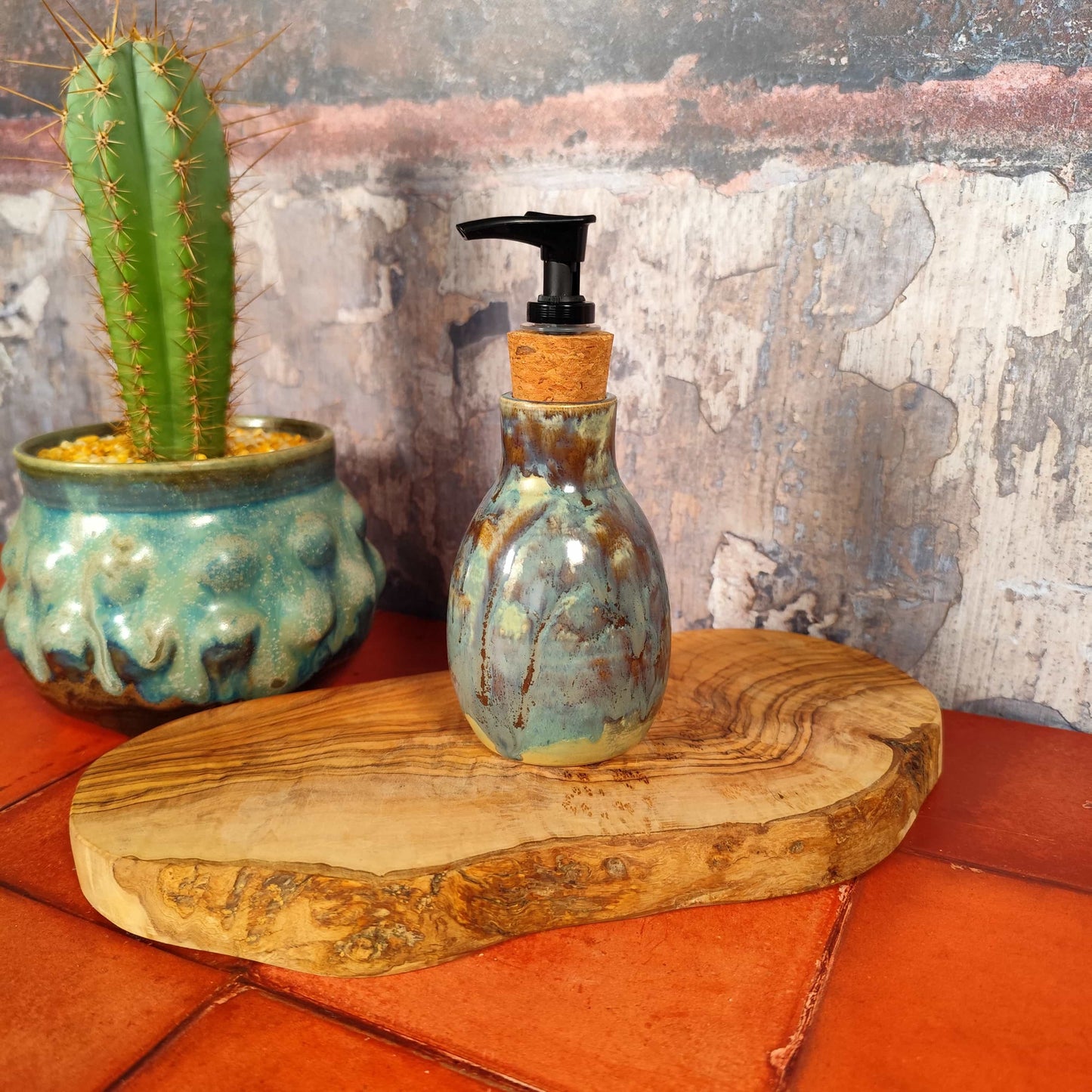 Unique glazed ceramic bathroom accessory gift idea. Ceramic soap dispenser in cerulean and olive glaze, made in Wales. On a wooden surface next to a cactus plant. 